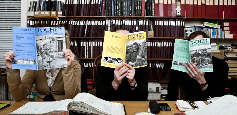 A row of three seated people who are facing the camera but their faces are obscured by issues of Tocher magazine, which they are holding up to read.