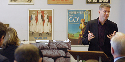 Photograph of man speaking to an audience in front of world war one memorabilia