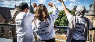 Three students in white shirts featuring the phrase "Challenge. Create. Change." stand side by side, waving outwardly from the rooftop of the Edinburgh Futures Institute.