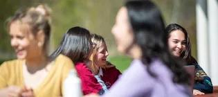 Groups of students gathered around tables, engaged in conversation in a pleasant outdoor environment.