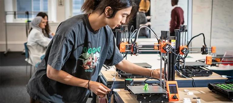 A student focused on operating a 3D printer in a modern classroom environment, surrounded by tools and printed objects.
