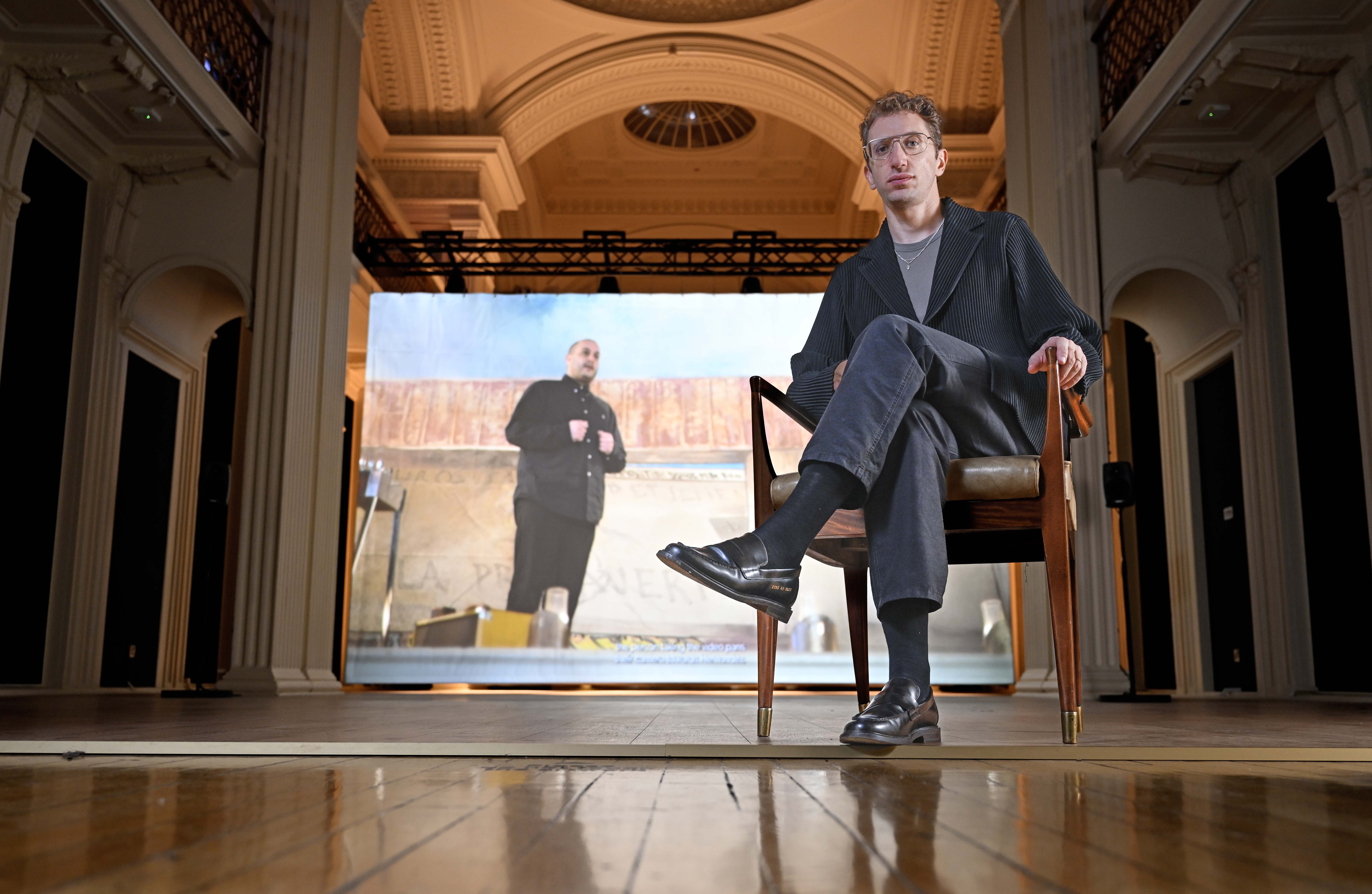 The artist sitting in front of a screen showcasing his work.